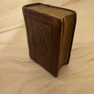 Puede incluir: Un libro antiguo, encuadernado en cuero, con un diseño en relieve detallado en la portada. La cubierta del libro es de color marrón oscuro, y las páginas tienen un tono dorado. El libro está ligeramente inclinado, mostrando su aspecto envejecido.