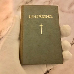 May include: A small, rectangular book with a faded teal cover. The gold text reads "IN HIS PRESENCE" above a gold cross. The book is held by a gloved hand, suggesting its delicate nature. The background is a marble-like surface.