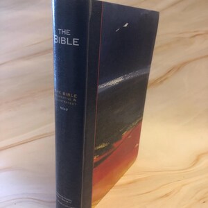 May include: A hardcover book titled "THE BIBLE" with a dark blue cover. The spine features the title in white lettering. The book's cover has a landscape illustration with red, orange, and black hues.