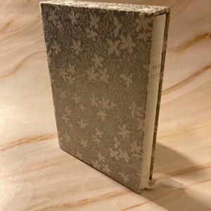 Könnte beinhalten: Ein graues Buch mit einem weißen Blumenmuster auf dem Einband. Das Buch ist geschlossen und steht aufrecht auf einer hellbraunen Oberfläche.