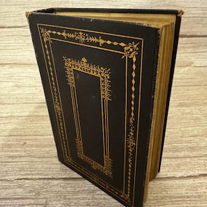 Peut inclure: Un livre relié vintage avec une couverture noire ornée de filigranes dorés complexes et de motifs géométriques. Le livre présente des tranches de pages jaunies par le temps, visibles sur le côté, témoignant de son ancienneté.