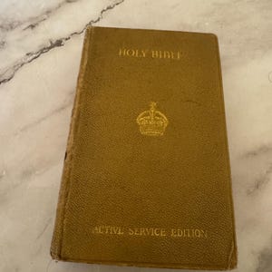 Peut inclure: Une Sainte Bible de couleur moutarde, édition Active Service. La couverture porte l'inscription dorée "Holy Bible" et "Active Service Edition". Un emblème doré est centré sous le titre. Le livre montre des signes d'usure.