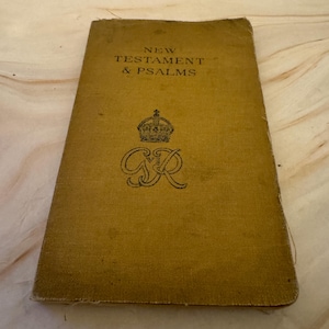 May include: A vintage book with a mustard-colored cover. The title "NEW TESTAMENT & PSALMS" is printed in black. A crown and the letters "GR" are also visible. The book appears to be old and well-loved.