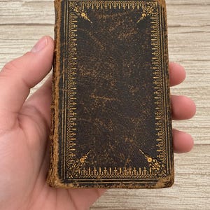 Puede incluir: Un libro antiguo, pequeño y rectangular, con una cubierta de cuero texturizado de color marrón oscuro. La portada presenta un intrincado borde decorativo estampado en oro con detalles florales en las esquinas y un patrón geométrico.
