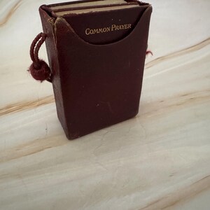 May include: A dark red leather book with the words "COMMON PRAYER" in gold lettering. The book is in a matching leather case with a knotted cord. The pages are visible, indicating it is a well-used item. The overall design is classic and compact.
