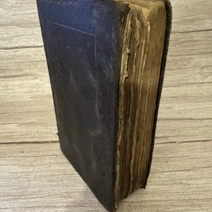 Puede incluir: Un libro antiguo con una cubierta de cuero oscuro y desgastado que muestra signos visibles de uso. Los bordes de la cubierta presentan detalles en relieve tenues, mientras que las páginas están envejecidas, amarillentas y tienen una textura rugosa.