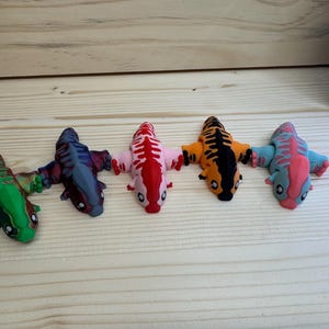 May include: Five small, colourful, 3D printed figurines of a creature with a long tail and a large head. The figurines are green, blue, red, orange, and pink.