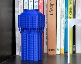 May include: A blue architectural model stands on a shelf with books. The model is a detailed replica of a building. A silver, arched shelf is in front of the books. The books have various titles and colors.