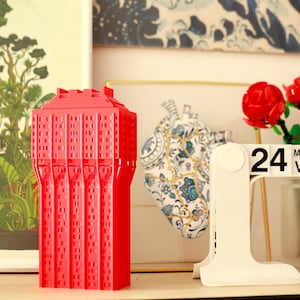 Könnte beinhalten: Ein rotes 3D-gedrucktes Modell eines Gebäudes, ein weißer Kalender mit dem Datum "24 MAG VEN" und ein Strauß roter Rosen.