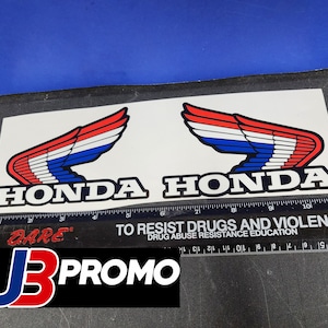 May include: A white sticker featuring the Honda logo, with two wing designs in red, white, and blue. The word "HONDA" is printed in black. A ruler and a "DARE" logo are also visible.