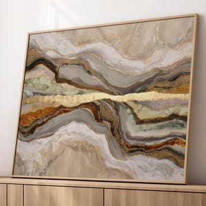 May include: Abstract art print with a natural landscape design. The artwork features a layered composition of beige, gold, brown, and grey tones, framed in a light wood. The piece is displayed on a wooden cabinet, with white vases and a round ottoman.