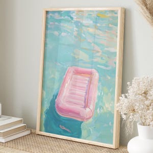 Könnte beinhalten: Ein gerahmtes Gemälde eines rosa aufblasbaren Floßes, das in einem türkisfarbenen Pool schwimmt. Das Gemälde hat einen hellen Holzrahmen.