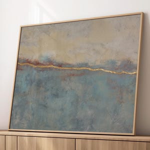 Puede incluir: Pintura abstracta de paisaje en un marco de madera. La obra presenta una superficie texturizada con una paleta de colores azul, beige y dorado. La pintura se exhibe sobre un mueble de madera, lo que sugiere un enfoque en la decoración del hogar y el arte mural.