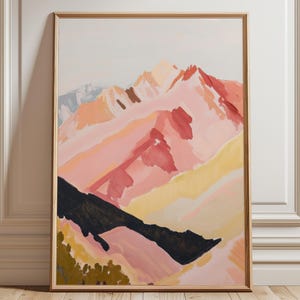 May include: A framed abstract mountain landscape painting. The artwork features pink, red, yellow, and black hues, with a light background. The painting is in a gold-coloured frame and leans against a white wall.