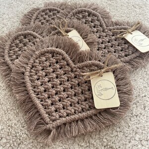 May include: Four brown heart-shaped crocheted coasters with fringe edges. Each coaster has a small, rectangular tag attached with the text "The Knot Craft Co."