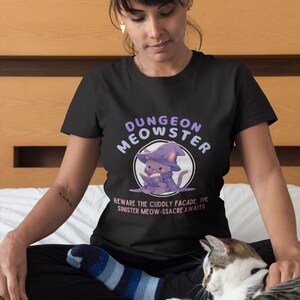 May include: A black t-shirt with a purple and white illustration of a cat wearing a wizard hat and holding a book. The text "Dungeon Meowster" is written in a purple font above the cat and the text "Beware the cuddly facade. The sinister meow-ssacre awaits" is written below the cat.
