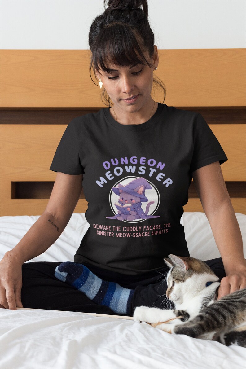 May include: A black t-shirt with a purple and white illustration of a cat wearing a wizard hat and holding a book. The text "Dungeon Meowster" is written in a purple font above the cat and the text "Beware the cuddly facade. The sinister meow-ssacre awaits" is written below the cat.