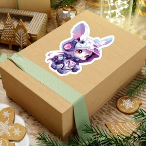 May include: A sticker featuring a cute anime-style character in a futuristic outfit with bunny ears. The character is primarily purple, white, and pink, with large orange eyes. The sticker is placed on a brown gift box with a green ribbon.