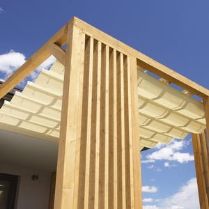 Op de afbeelding: Houten pergola met een intrekbaar crèmekleurig zonnescherm, tegen een helderblauwe lucht met verspreide wolken. De pergola heeft verticale houten lamellen en een stevig frame, dat schaduw en architectonische interesse biedt.