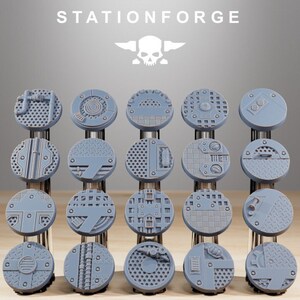 Peut inclure: Une collection de socles miniatures circulaires gris avec divers motifs industriels. Les socles sont disposés en rangées et en colonnes, présentant différentes textures et motifs. Le texte "STATIONFORGE" et un logo de crâne sont visibles en haut.