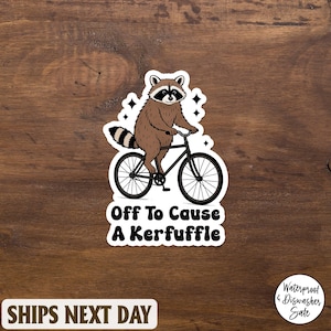 Puede incluir: Una pegatina con un mapache montando en bicicleta. El mapache es marrón y negro con una cola a rayas. El texto "Off To Cause A Kerfuffle" está debajo. La pegatina está sobre un fondo de madera.