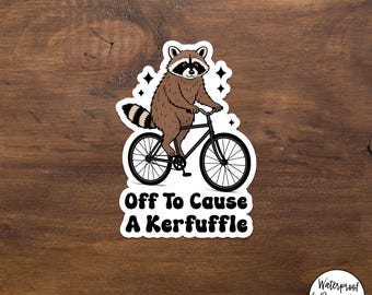 May include: A sticker featuring a raccoon riding a bicycle. The raccoon is brown and black with a striped tail. The text "Off To Cause A Kerfuffle" is below the raccoon. The sticker is on a wooden background.