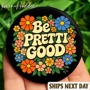 Pode incluir: Um botão circular preto com as palavras "Be PRETTI GOOD" em amarelo, rodeado por flores coloridas em laranja, rosa e azul. O botão tem um design retro.