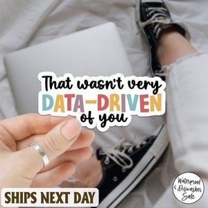 Könnte beinhalten: Ein farbenfroher Aufkleber mit dem Text "That wasn't very DATA-DRIVEN of you." Der Aufkleber hat einen weißen Hintergrund und wird von einer Hand mit einem silbernen Ring gehalten. Ein Laptop, schwarze Turnschuhe und eine weiße Hose sind im Hintergrund.