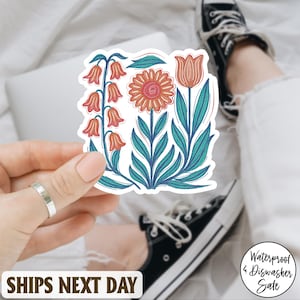 May include: A floral sticker featuring stylized orange and pink flowers with teal leaves. The sticker is held in a hand, with a laptop and a person's legs in the background. The sticker is waterproof and dishwasher safe.
