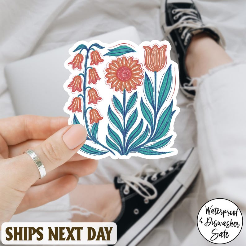May include: A floral sticker featuring stylized orange and pink flowers with teal leaves. The sticker is held in a hand, with a laptop and a person's legs in the background. The sticker is waterproof and dishwasher safe.