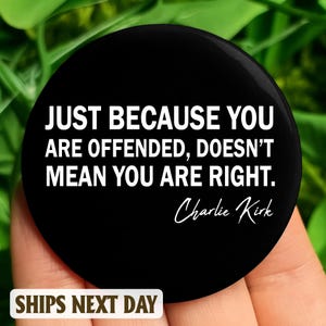 Op de afbeelding: Een zwarte ronde button met witte tekst: "JUST BECAUSE YOU ARE OFFENDED, DOESN'T MEAN YOU ARE RIGHT." Toegeschreven aan Charlie Kirk. De button wordt tegen een groene achtergrond gehouden.