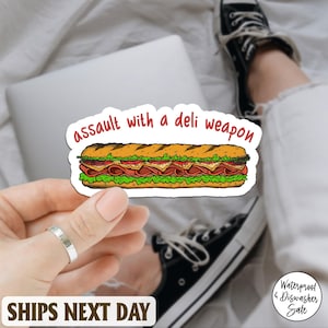 May include: A sticker featuring a cartoon submarine sandwich with the text "assault with a deli weapon" in red. The sandwich has layers of meat, cheese, and lettuce. The sticker is held by a hand.