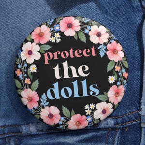 May include: A black button with a floral wreath border. The text "protect the dolls" is written in pink, white, and blue lettering.