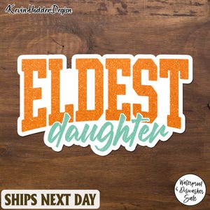 Op de afbeelding: Een sticker met de woorden "ELDEST daughter" in oranje glitterletters met een witte rand. Het woord "daughter" is in een turquoise scriptlettertype. De sticker ligt op een bruin houten oppervlak.