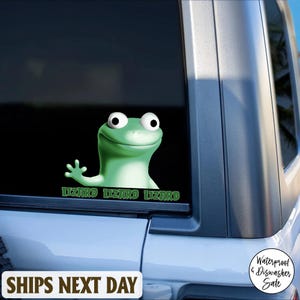 May include: A green lizard sticker with the word "LIZARD" repeated three times in green text. The sticker is on a car window. The lizard has large white eyes and is waving. The sticker is waterproof and dishwasher safe.