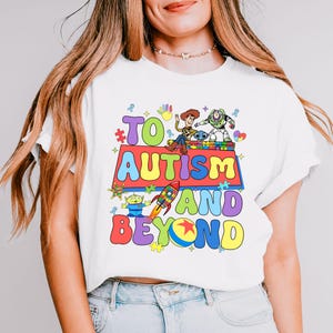 Könnte beinhalten: Weißes T-Shirt mit einer bunten Grafik mit Figuren aus dem Toy Story-Franchise. Die Grafik lautet "#TO AUTISM AND BEYOND" mit Puzzle-Teilen und anderen bunten Designs.