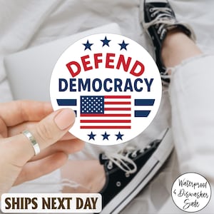 May include: A round white sticker with the words "DEFEND DEMOCRACY" in red and blue, featuring the American flag. The sticker is held by a hand with a silver ring. The background includes a laptop, white pants, and black sneakers.