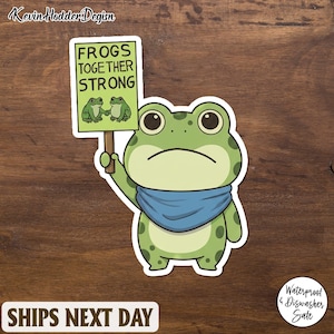 May include: A green frog sticker with a blue scarf and a sign that reads "Frogs Together Strong." The sticker features a cartoon frog holding a sign with two frogs on it. The sticker is waterproof and dishwasher safe.