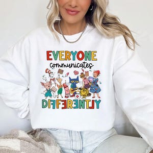 Puede incluir: Sudadera blanca con un gráfico colorido de personajes de libros infantiles. El texto dice "Todos nos comunicamos de manera diferente".