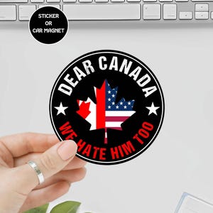 May include: A round sticker or car magnet with the text "DEAR CANADA" and "WE HATE HIM TOO" in white. The design features a maple leaf with the colors of the American and Canadian flags.