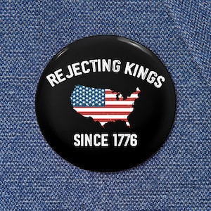 Puede incluir: Un botón redondo negro con el texto "Rejecting Kings" en blanco, arqueado en la parte superior. El botón presenta un mapa de los Estados Unidos con un diseño de bandera y el texto "Since 1776" en blanco.