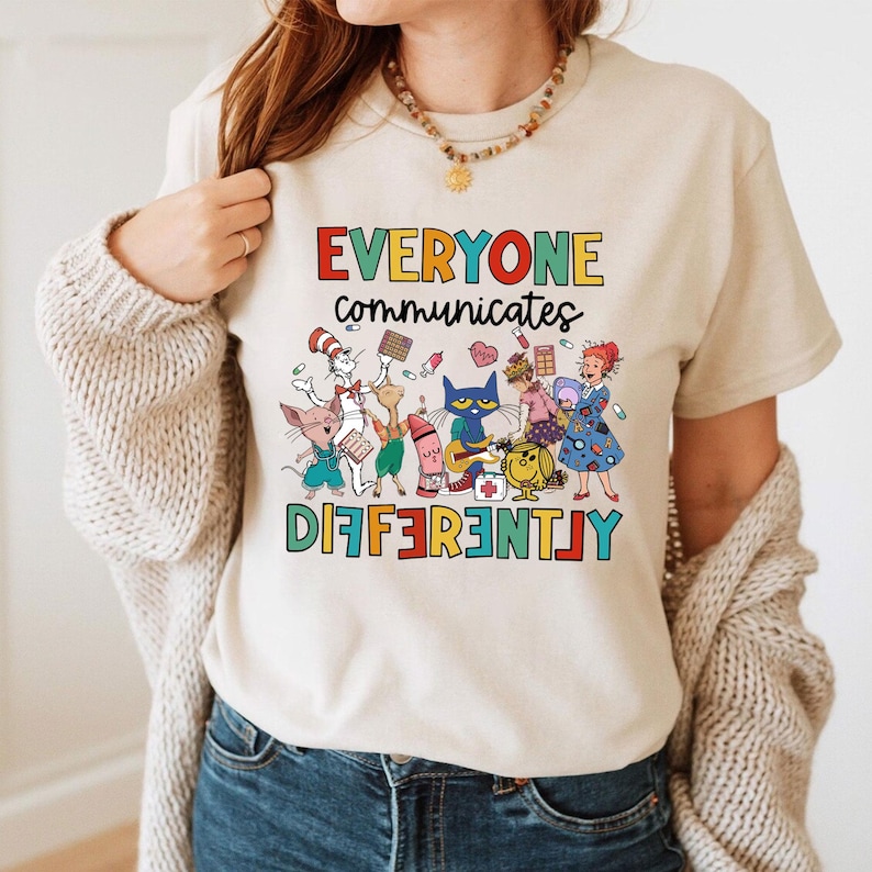 以下が含まれることがあります： ベージュのTシャツに、カラフルな漫画のキャラクターと「みんな違う方法でコミュニケーションをとる」というテキストが描かれています。