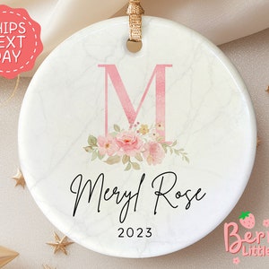May include: A white ceramic ornament with a pink marble design. The ornament features a large pink letter "M" with pink floral accents and the text "Meryl Rose 2023".