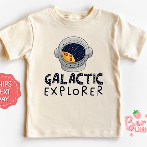 May include: A cream colored t-shirt with a cartoon astronaut wearing a helmet with a pizza moon inside. The text "GALACTIC EXPLORER" is printed below the astronaut.