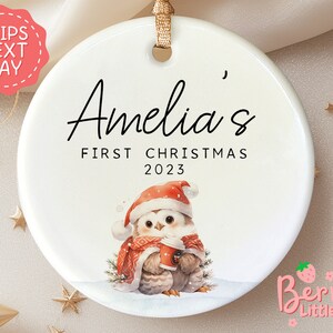 May include: White ceramic Christmas ornament with a cute cartoon owl wearing a Santa hat and holding a cup of hot chocolate. The ornament is personalized with the text "Amelia's First Christmas 2023".