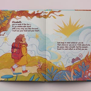Op de afbeelding: Een ge&iuml;llustreerd boek met een kleurrijke omslag met een jonge persoon met een rugzak die op een pad loopt. De tekst op de pagina luidt "Elizabeth. you've made it this far- It's an unbelievable start! with every step you take forward You'll use your head and your heart. Just keep in mind whatever you do That wherever you are is YOUR place to be. So pause, take a breath, feel the moment... Soak it in and reflect on all that you see."