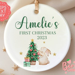 May include: A white ceramic Christmas ornament with the text "Amelie's First Christmas 2023" in green script. The ornament features a watercolor illustration of a Christmas tree, a bunny, gifts, and gold stars. A gold ribbon is attached for hanging.