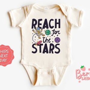 May include: A cream-colored baby onesie with a black graphic of an astronaut reaching for the stars. The text "Reach for the Stars" is printed in black.