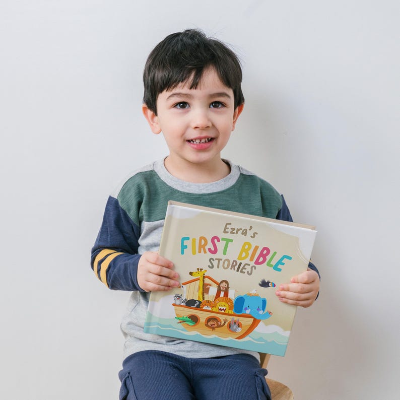 May include: A young child holding a book titled "Ezra's First Bible Stories." The book cover features a colorful illustration of Noah's Ark with various animals. The child is wearing a long-sleeved shirt with green, gray, and navy blue panels.