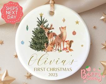 May include: White ceramic Christmas ornament with a watercolor illustration of a forest scene. The scene includes a Christmas tree, a deer, a fox, a squirrel, and a bird. The ornament is personalized with the name "Olivia's" and the text "First Christmas 2023".